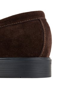 Brązowy zamszowy but z zaokrąglonym obcasem, z wyraźnym przeszyciem i teksturowaną czarną gumową podeszwą. Zbliżenie podkreśla detale materiału i designu.