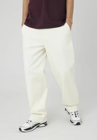 Pantalons en coton blancs avec une coupe ample, dotés de poches latérales et d'une texture lisse, associés à des chaussures de sport noires et argentées.