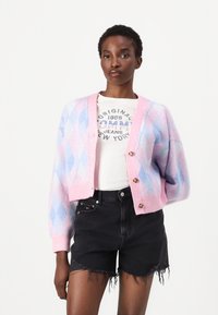 Cárdigan de rombos en tonos rosa pastel y azul, con un diseño corto, que cuenta con un cierre de botones y puños acanalados, llevado sobre una camiseta gráfica blanca y pantalones cortos negros.
