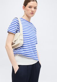 Γυναίκα που φορά μπλούζα με μπλε και λευκές ρίγες και μαύρο παντελόνι, κρατώντας μπεζ τσάντα ώμου με τα χέρια στις τσέπες.