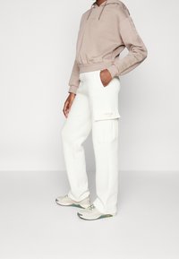 Beige hoodie med snøre og logo, parret med hvide cargo-bukser med en klap lomme. Lyse sneakers med grønne detaljer.