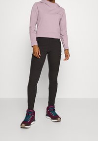 Světlé fialové cropped hoodie s širokým výstřihem a logem, kombinované s černými zúženými kalhotami s texturovanými akcenty a zipovými kapsami, obuté s burgundskými teniskami.