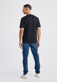 Camiseta de algodón negra de mangas cortas, combinada con jeans de mezclilla azules y zapatillas blancas, vista desde la parte trasera.