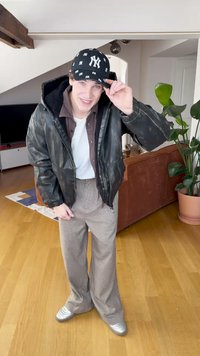 Melna ādas bomber žakete, brūna krekls, balta T-krekls, rakstainas plašas bikses un sudraba apavi ar Ņujorkas beisbola cepuri.