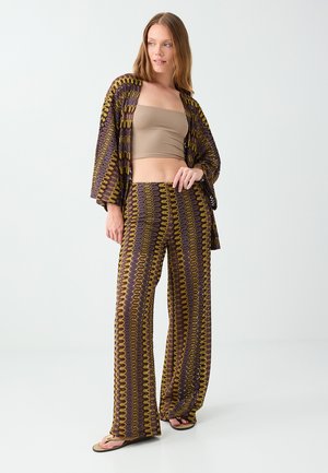 Donna che indossa un crop top marrone, pantaloni ampi a fantasia e un cardigan aperto abbinato con infradito, in piedi davanti a uno sfondo semplice.