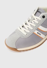 Grauer Stoff- und Wildledersneaker mit weißen und orangen Streifen, weißen Schnürsenkeln und einer Gummisohle. Auf der Außenseite befindet sich ein Logo.