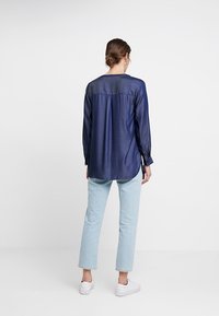 Camicia di denim a maniche lunghe, colore blu scuro, texture liscia, presenta un dettaglio con bottoni sul retro, abbinata a jeans azzurri e sneaker bianche.