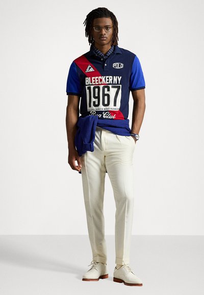 Tengerészkék póló piros és fehér kiegészítőkkel, nagy nyomtatott felirattal: "BLEECKER NY 1967." Krém színű nadrág és világos színű cipők teszik teljessé a megjelenést.
