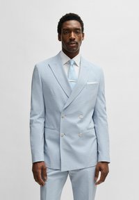 Ljusblå dubbelknäppt kostym i strukturerad tyg, med slag i ände, vit skjorta och ljusblå slips med metallaccent.