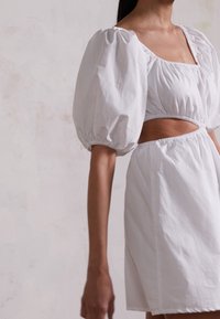 Vit kort klänning med puffärmar och fyrkantig halsringning, med en öppning vid midjan och samlad elastisk detalj för en figurnära look.