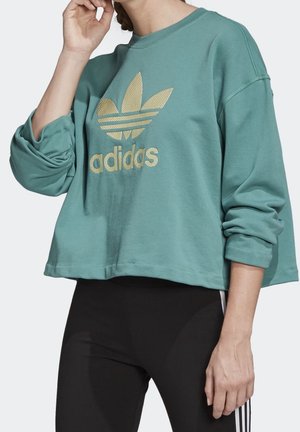 Osoba v tyrkysové mikině Adidas se zlatým logem a černých legínách s bílými pruhy, ruka u obličeje, uvolněná póza.