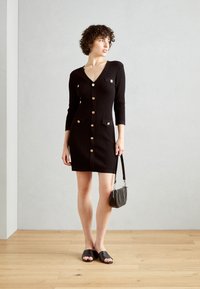 Robe tricotée noire avec un décolleté en V, des manches trois-quarts, des boutons dorés et des poches avant. Associée à des sandales noires à enfiler et un petit sac.