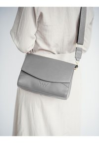 Šedá kožená taška přes rameno s klopovým uzávěrem, s vyraženým logem, nastavitelným popruhem a hladkým povrchem. Minimalistický design.