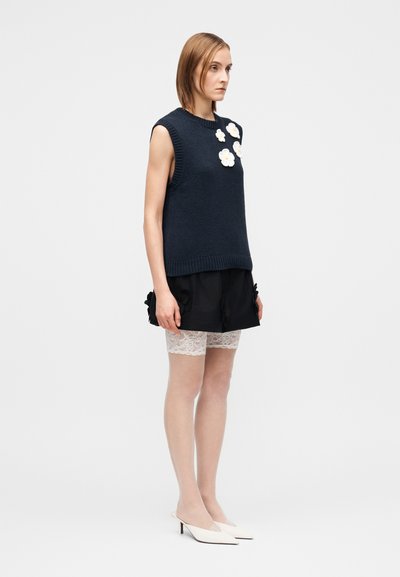 Marineblauer ärmelloser Strickpullover mit Blumen-Applikationen, kombiniert mit schwarzen Shorts mit Rüschen und weißen Spitzenstrumpfhosen, abgerundet mit weißen, hochhackigen Mules.
