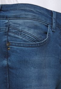 Jeans de mezclilla azul oscuro con un diseño ajustado, costuras visibles y dos botones decorativos en el bolsillo trasero. La superficie tiene una textura que muestra un ligero desgaste.