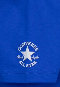 Tela de algodón azul con un logotipo circular blanco que incluye una estrella y el texto "CONVERSE Chuck Taylor ALL STAR" en una fuente en negrita.