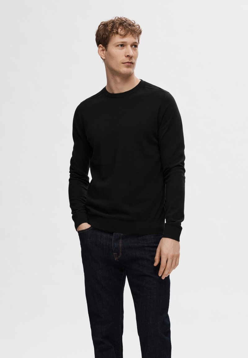Sweater preto com decote redondo e mangas longas, feito de um tecido liso, combinado com jeans escuros de denim, design minimalista, sem padrões visíveis.