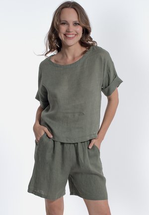 Donna sorridente con le mani nelle tasche, indossa una maglia verde oliva a maniche corte larga e pantaloncini coordinati su uno sfondo bianco semplice.