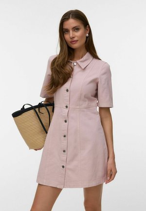 Mujer con un vestido de botones en la parte delantera de color rosa pálido y mangas cortas, sosteniendo un bolso de paja con asas negras, sobre un fondo blanco liso.
