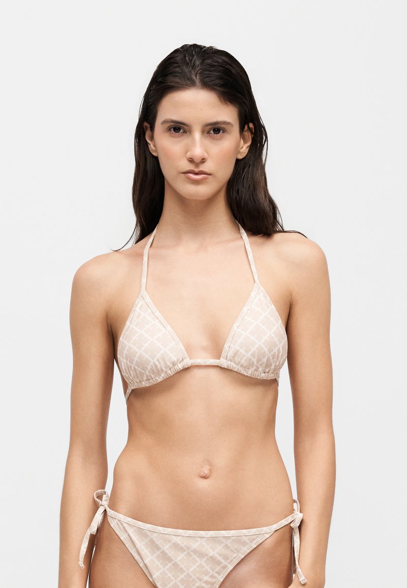 Femme aux longs cheveux foncés portant un haut de bikini triangle beige clair à motifs et un bas de bikini assorti à nouer sur les côtés sur un fond uni.