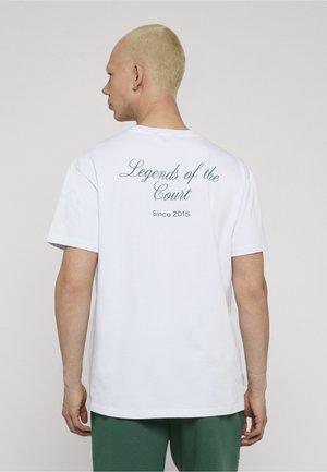 Person med kort blondt hår iført en hvit T-skjorte med teksten "Legends of the Court Since 2015" på baksiden, kombinert med grønne bukser.