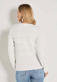 Frau mit blonden Haaren, die einen weißen langärmligen Strickpullover mit subtilen horizontalen Löchern und hellblauen Jeans trägt, steht mit dem Rücken zugewandt.