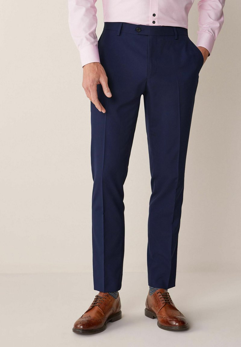 Pantalones navy de corte entallado, con pliegue frontal, bolsillos laterales y una textura de tela suave, combinados con zapatos de vestir marrones.