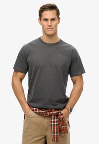 Maglietta di cotone grigia con maniche corte e collo tondo, con un piccolo logo. Abbinata a pantaloni khaki e una camicia a quadri legata in vita.