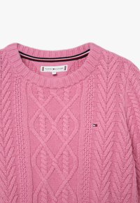 Rosa kabelstickad tröja med rund halsringning som har en liten Tommy Hilfiger-logotyp på bröstet och en märkskylt inuti kragen.