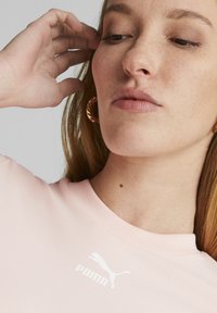 Lichtroze shirt met korte mouwen met een wit Puma-logo. Close-upweergave, gladde textuur en ronde halslijn, met een hand die het haar aanraakt.