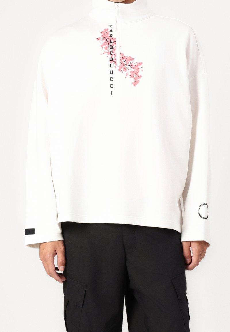 Pull blanc à manches longues avec fleur de cerisier rose et texte noir vertical, demi-fermeture éclair devant, porté avec un pantalon cargo noir.