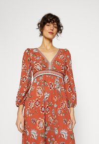 Vestido maxi floral em ferrugem com decote em V, mangas bufantes e cintura ajustada. Apresenta um tecido texturizado com estampas florais vibrantes em azul e rosa.
