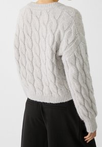 Hellgrauer Kabelstrickpullover mit lockerer Passform, geripptem Rundhalskragen und Bündchenärmeln, strukturierter Oberfläche, getragen zu schwarzen Hosen.