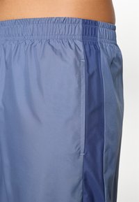 Shorts de sport bleus avec une taille élastique, présentant une texture lisse, une poche latérale et un panneau contrastant bleu foncé sur le côté gauche.