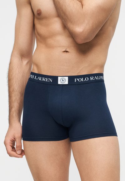 Torso masculin portant un boxer Polo Ralph Lauren bleu marine avec le logo sur la ceinture, sur un fond clair uni.
