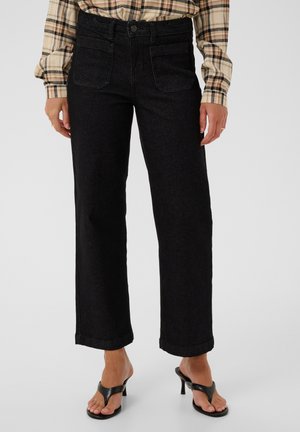 Person iført sort, højtaljede jeans med lige ben og forlommer, en beige ternet skjorte og sorte sandaletter med hæl.