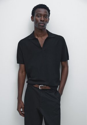 Jeune homme portant une chemise noire à manches courtes avec col et un pantalon noir ceinturé de marron, debout devant un fond clair uni.