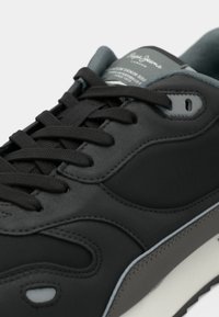 Zapatilla negra con parte superior sintética lisa, acentos grises, cuello acolchado, puntera redonda y cordones texturizados; presenta una etiqueta con el logotipo en la lengüeta.