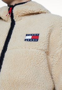 Kremowa polarowa kurtka z czarnym zamkiem błyskawicznym i czerwonym paskiem do ściągania. Posiada naszywkę z logo Tommy Jeans na piersi.