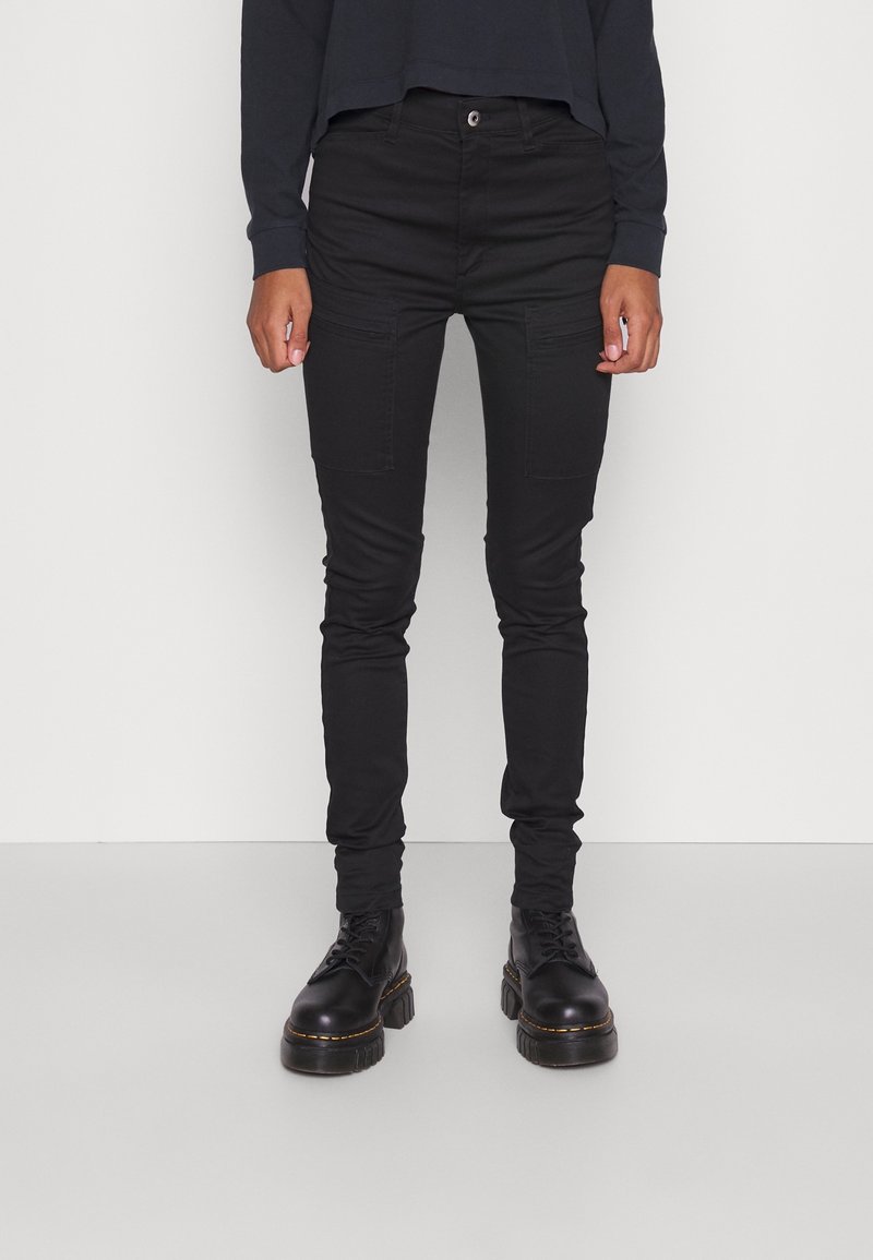 Personne portant un pantalon cargo slim noir avec des poches plaquées à l'avant et des bottes à plateforme noires lacées, debout devant un fond uni.