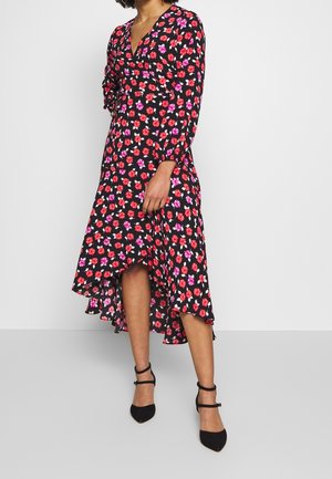 Woman wearing black long-sleeve floral dress with red and purple flowers, asymmetrical hem, and black ankle-strap heels, standing against white wall.