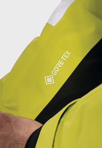 Giacca GORE-TEX con un esterno di un brillante giallo neon, dettagli neri e un logo bianco. Presenta una texture liscia e impermeabile.