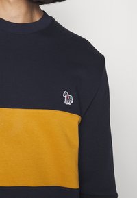 Maglietta a blocchi di colore blu navy e senape con piccola zebra multicolore ricamata sul petto, indossata da una persona.