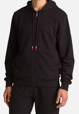 Mies, joka pitää mustaa vetoketjullista hupullista hupparia, jossa on punaiset, valkoiset ja siniset nyörit, yhdistettynä mustiin housuihin.