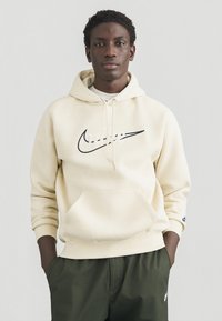 Kremasta kapuca z mornarsko modrim logotipom Nike, z žepom spredaj in vrvicami. Oblečena s temno zelenimi hlačami, na svetlem ozadju.