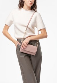 Borsa a tracolla in pelle rosa con pattina pieghevole, tracolla in catena dorata e texture liscia. Indossata con un maglione chiaro e pantaloni grigi.