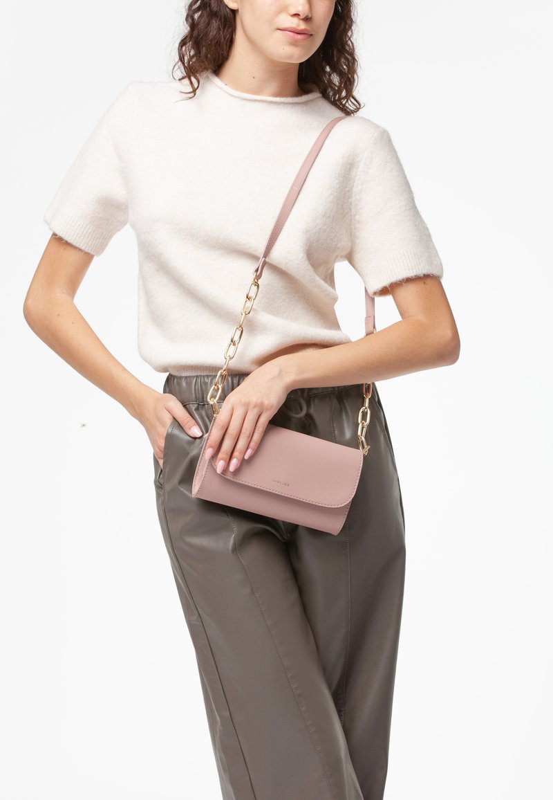 Borsa a tracolla in pelle rosa con pattina pieghevole, tracolla in catena dorata e texture liscia. Indossata con un maglione chiaro e pantaloni grigi.