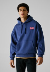Sudadera azul marino de tejido suave, con un bolsillo frontal y un parche del logo "Red Bull Racing" en el pecho. Puños y dobladillo acanalados.