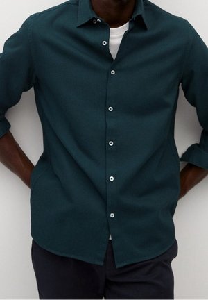 Mann trägt ein dunkelgrünes Hemd über einem weißen Unterhemd, die Ärmel hochgekrempelt, die Hände auf der Hüfte ruhend.