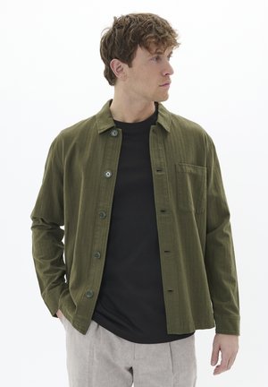 Olivgrünes, lässig geschnittenes Hemd mit strukturiertem Stoff und einer Brusttasche, getragen über einem schwarzen T-Shirt und hellen Hosen.
